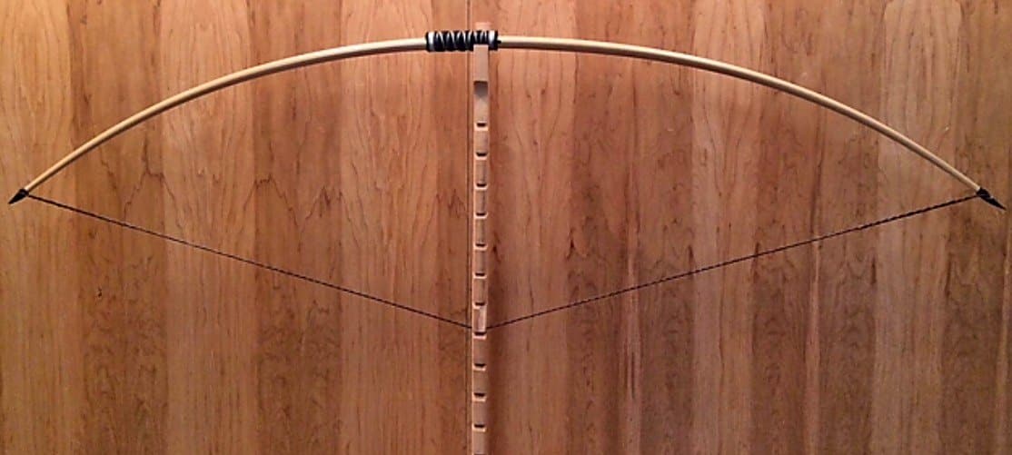 A pale longbow drawn to 22 inches on Doug's tiller against the maple wall